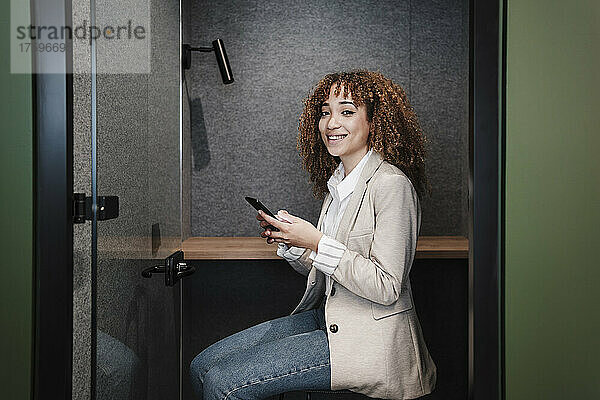 Smiling young businesswoman with smart phone sitting at desk in cabin