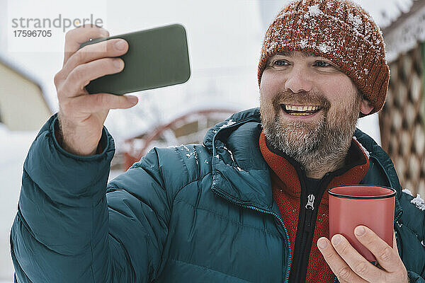Lächelnder Mann hält Kaffeetasse in der Hand und macht im Winter ein Selfie mit dem Handy
