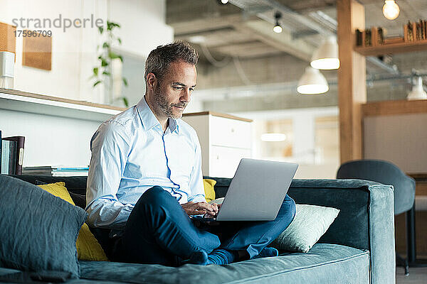 Geschäftsmann sitzt mit gekreuzten Beinen auf Sofa im Büro und benutzt Laptop