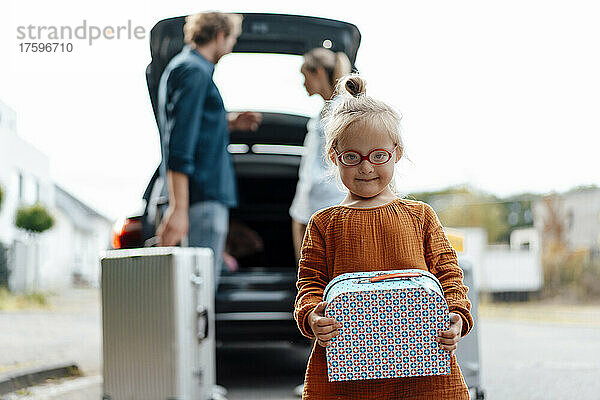 Mädchen hält Koffer in der Hand  Eltern laden Gepäck ins Auto