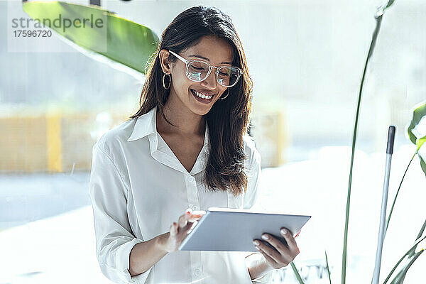 Lächelnde junge Geschäftsfrau mit Tablet-PC vor dem Fenster an einem sonnigen Tag