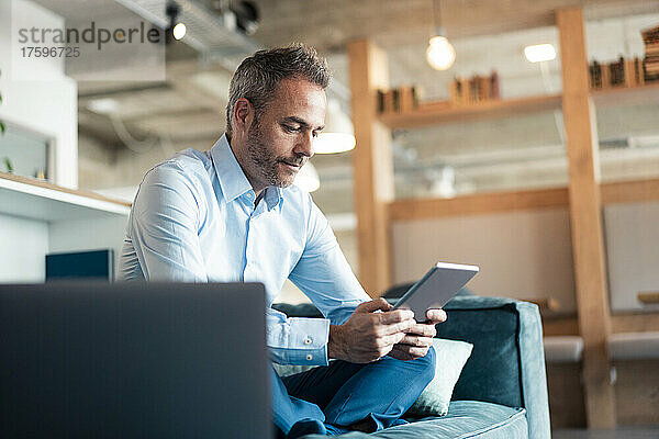 Businessman using tablet PC sitting cross-legged on sofa at office