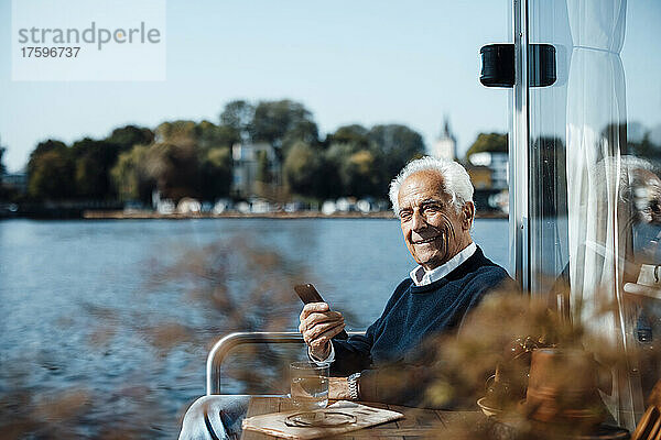 Lächelnder älterer Mann mit Smartphone sitzt an einem sonnigen Tag am Hausboot