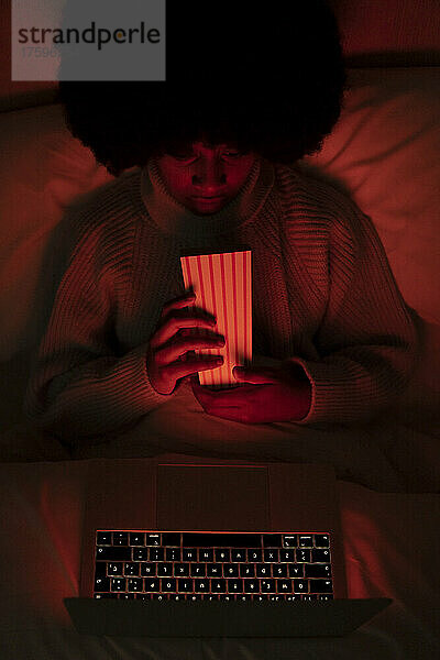 Afro-Frau mit Popcornbehälter schaut auf Laptop im Bett zu