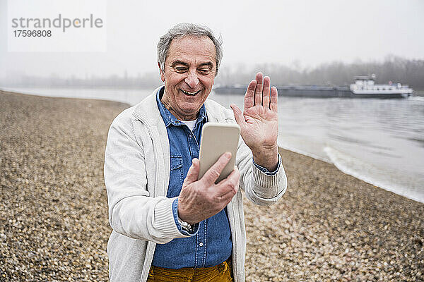 Glücklicher älterer Mann  der am Strand einen Videoanruf über sein Smartphone macht und mit der Hand winkt