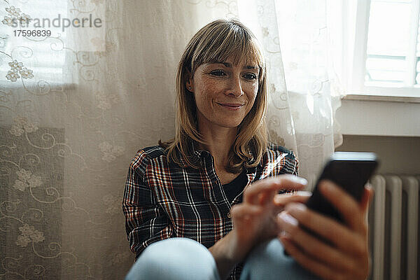 Lächelnde Frau benutzt Smartphone am Fenster zu Hause