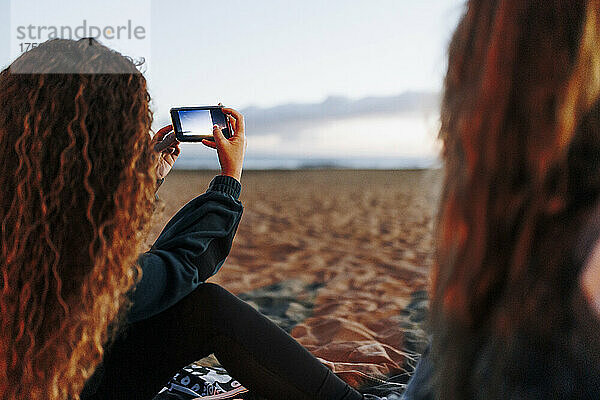 Junge Frau fotografiert mit Smartphone von Freund am Strand