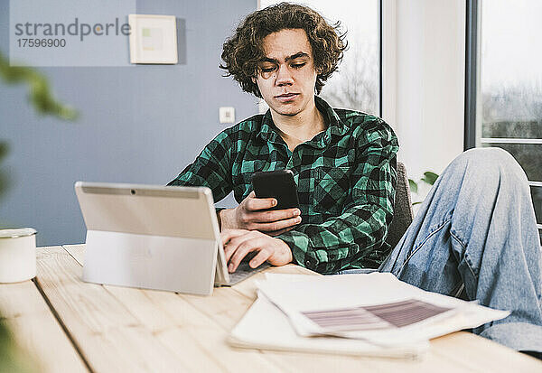 Junge Studentin sitzt mit Tablet-PC und nutzt Mobiltelefon am Tisch im Wohnzimmer