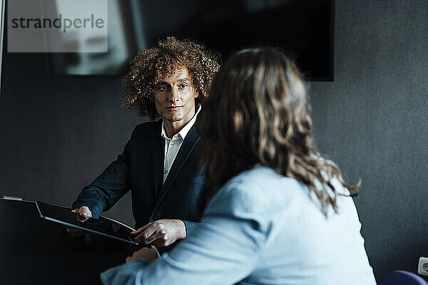 Geschäftsmann mit lockigem Haar hält einen Tablet-PC in der Hand und blickt einem Kollegen zu  der im Coworking-Büro diskutiert
