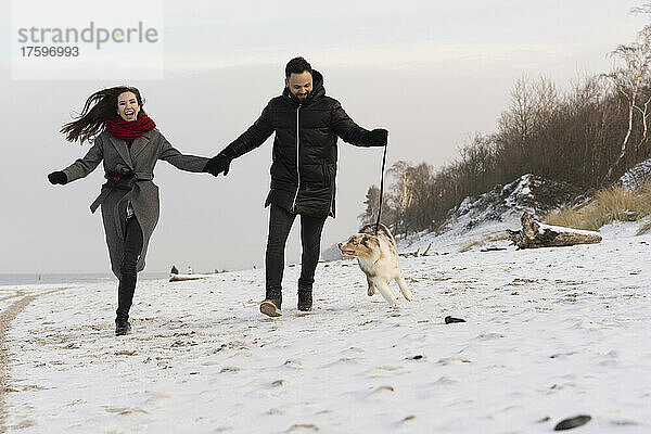 Glückliches junges Paar läuft mit Hund bei Sonnenuntergang im Schnee