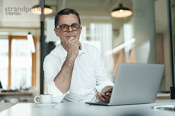 Selbstbewusster Geschäftsmann mit der Hand am Kinn sitzt mit Laptop auf dem Tisch im Büro