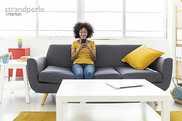 Lächelnde junge Frau  die zu Hause auf dem Sofa im Wohnzimmer sitzt und ihr Smartphone benutzt