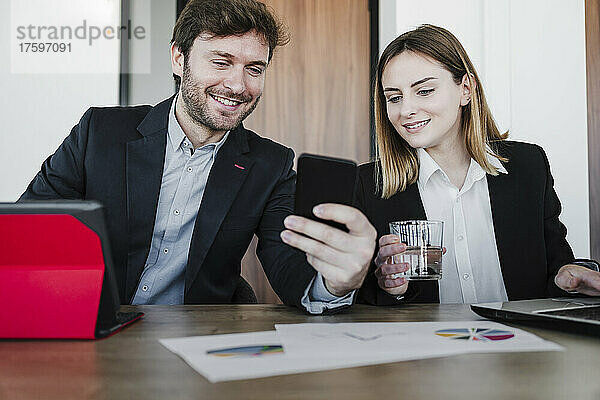 Lächelnde Geschäftskollegen teilen ihr Smartphone am Schreibtisch im Coworking-Büro