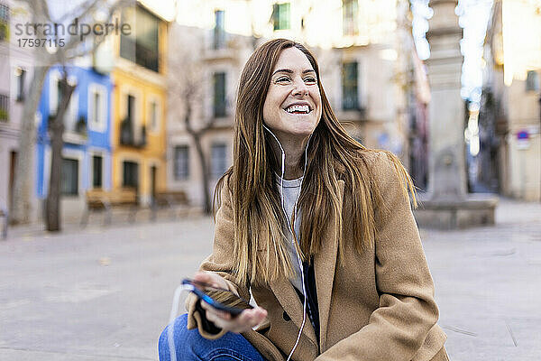 Glückliche Frau  die auf der Stadtstraße über In-Ear-Kopfhörer mit dem Smartphone spricht
