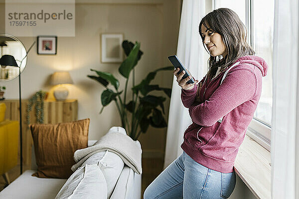 Frau schreibt Textnachrichten über Smartphone am Fenster zu Hause
