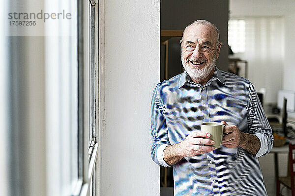 Glücklicher leitender Freiberufler hält eine Kaffeetasse in der Hand und blickt durch das Fenster