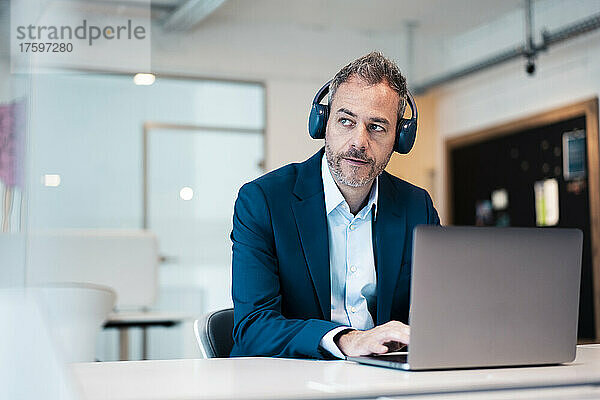 Nachdenklicher Geschäftsmann mit kabellosen Kopfhörern und Laptop am Schreibtisch am Arbeitsplatz