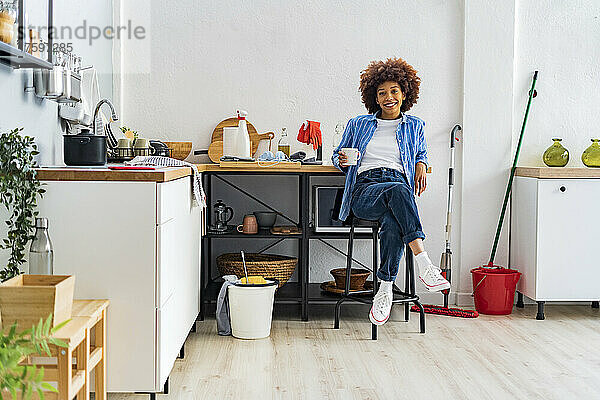 Lächelnde junge Afro-Frau sitzt auf einem Hocker und lehnt an der Küchentheke und entspannt sich zu Hause