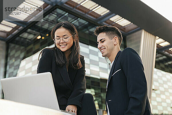 Lächelnde Geschäftskollegen arbeiten am Laptop vor dem Bürogebäude