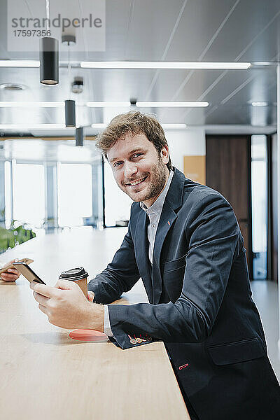 Glücklicher Geschäftsmann mit Mobiltelefon und Einwegkaffeetasse sitzt am Schreibtisch im Büro