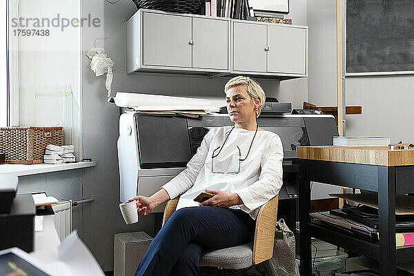 Nachdenkliche blonde Frau hält einen Becher in der Hand und sitzt auf einem Stuhl am Arbeitsplatz