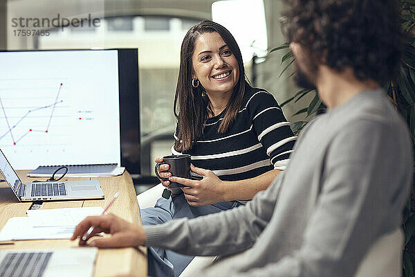 Lächelnde Geschäftsfrau hält Kaffeetasse in der Hand und unterhält sich mit einem Geschäftsmann  der am Konferenztisch sitzt