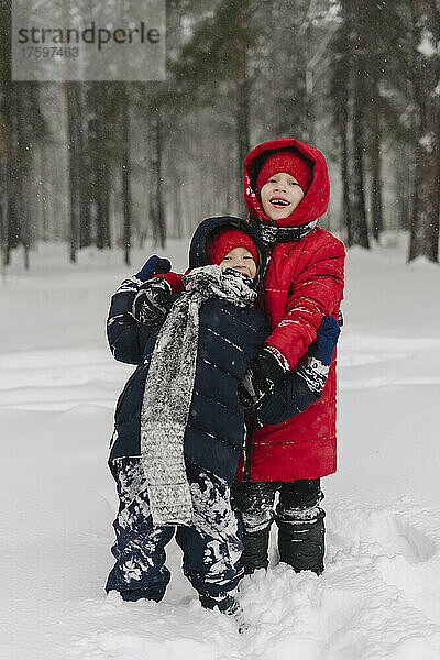 Lächelnder Junge umarmt Bruder  der auf Schnee im Wald steht