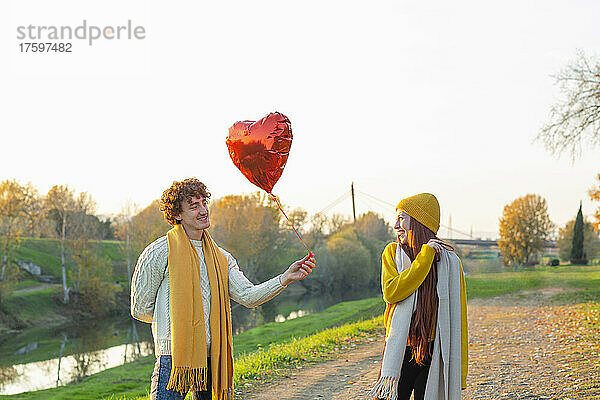 Smiling young man giving red heart shape balloon to cheerful girlfriend at autumn park