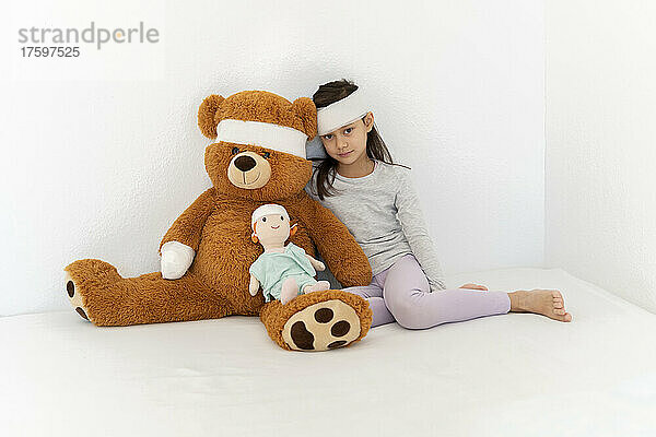 Bandage on girl with teddy bear and doll sitting in front of white wall