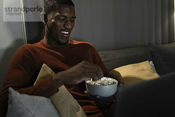 Smiling man watching movie on laptop eating popcorn at home