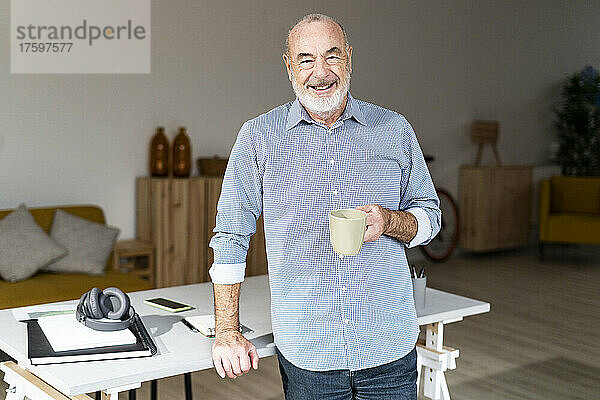 Glücklicher Geschäftsmann hält zu Hause eine Kaffeetasse am Schreibtisch