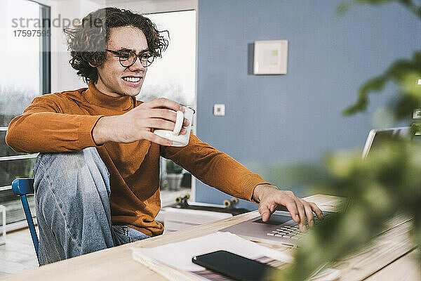 Glücklicher junger Mann  der zu Hause eine Kaffeetasse mit einem Laptop in der Hand hält