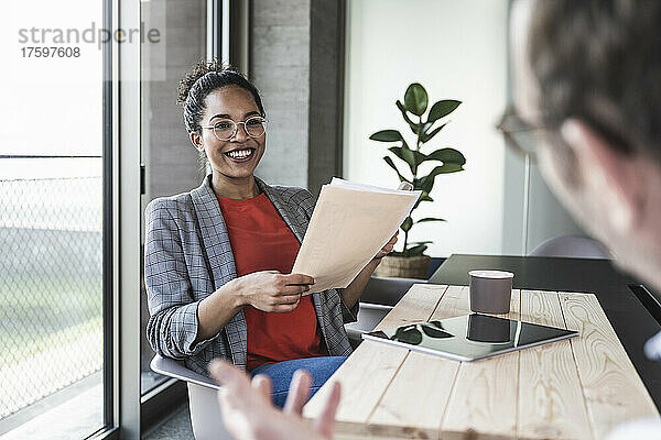 Lächelnde Geschäftsfrau hält Dokumente in der Hand und diskutiert mit einem Kollegen im Büro