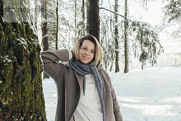 Lächelnde Frau steht mit den Händen hinter dem Kopf am Baum im Winterwald