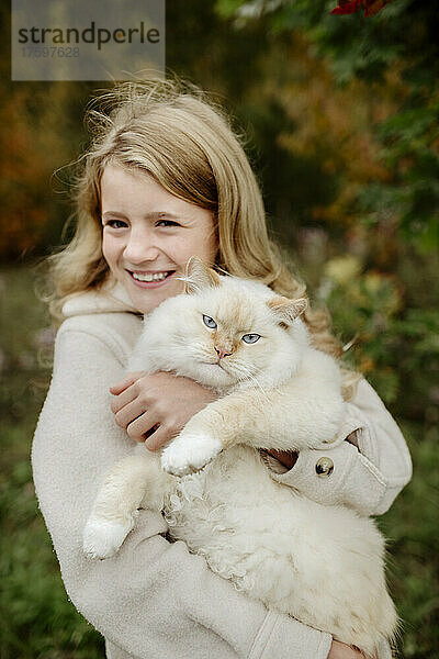 Glückliches blondes Mädchen mit Ragdoll-Katze im Garten