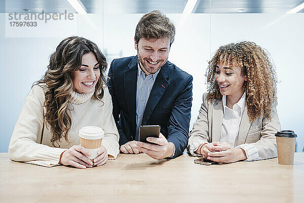 Glücklicher Geschäftsmann  der sein Smartphone mit Geschäftsfrauen teilt  die am Schreibtisch sitzen