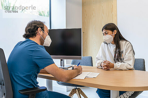 Arzt diskutiert mit Patient  der während einer Pandemie am Schreibtisch im Krankenhaus sitzt