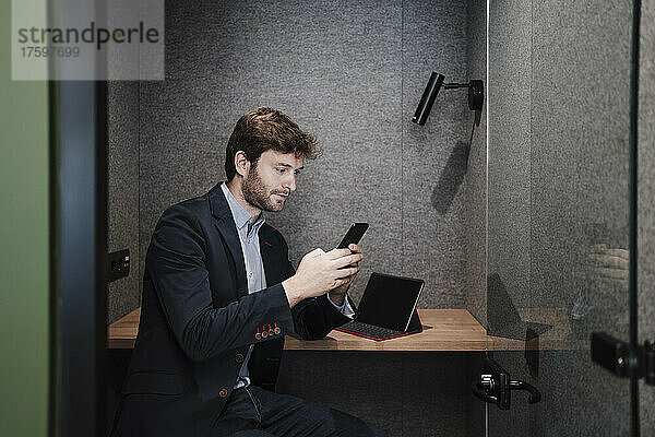 Geschäftsmann sitzt am Schreibtisch in der Kabine und benutzt Smartphone