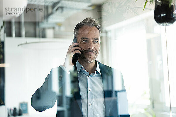 Lächelnder Geschäftsmann  der durch die Glaswand wegschaut und mit dem Mobiltelefon telefoniert