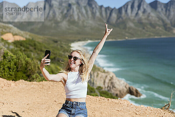 Glückliche Frau gestikuliert Friedenszeichen und macht an einem sonnigen Tag ein Selfie mit dem Handy