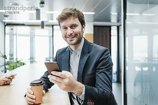 Lächelnder Geschäftsmann mit Einwegkaffeetasse und Smartphone am Schreibtisch