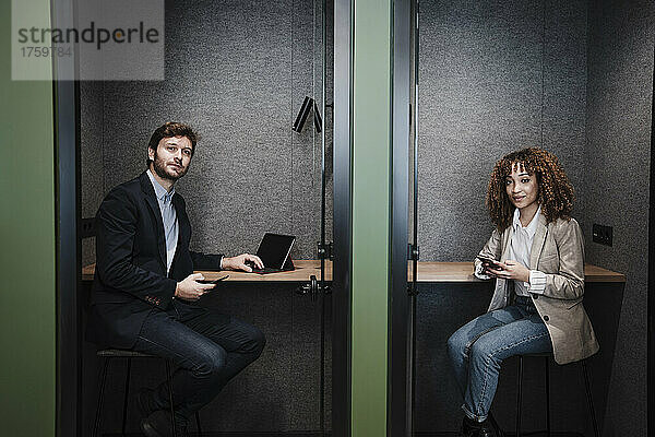 Geschäftsfrau und Geschäftsmann mit Mobiltelefonen sitzen am Schreibtisch in der Kabine