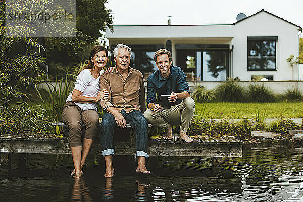 Lächelnder Mann mit Tablet-PC neben Vater und Mutter  der auf dem Steg am See im Garten sitzt