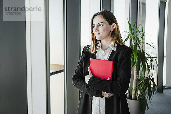 Lächelnde Geschäftsfrau mit Tablet-PC steht am Fenster im Büro