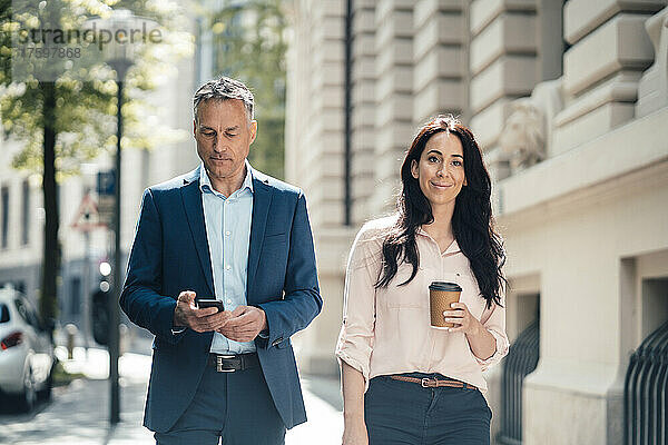 Geschäftsfrau mit Einwegkaffeetasse läuft an Geschäftsmann vorbei  der sein Smartphone auf dem Fußweg nutzt