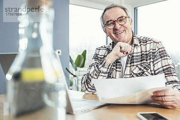 Glücklicher älterer Mann mit Brille sitzt mit Finanzrechnung am Tisch