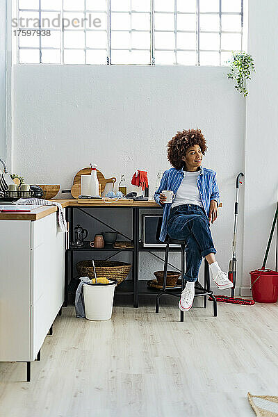 Nachdenkliche junge Afro-Frau sitzt auf einem Hocker und lehnt an der Küchentheke und entspannt sich zu Hause
