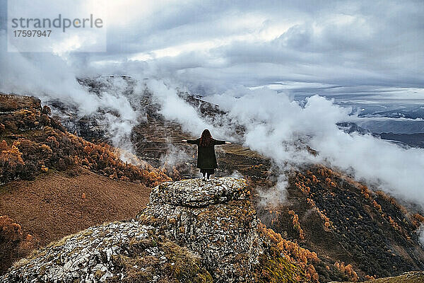 Female hiker standing with outstretched arms on top of outcrop in autumn mountains of North Caucasus