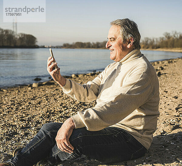 Glücklicher älterer Mann  der an einem sonnigen Tag am Strand einen Videoanruf über sein Mobiltelefon tätigt