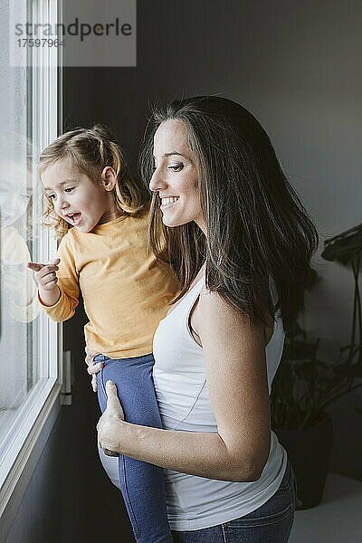 Glückliche schwangere Frau mit Tochter  die zu Hause durch das Fenster schaut